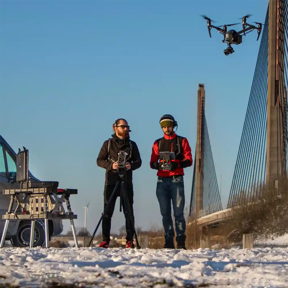 A professional two-person drone crew from Zero Gravity operating a cinema drone in a snowy landscape near a large cable-stayed bridge.